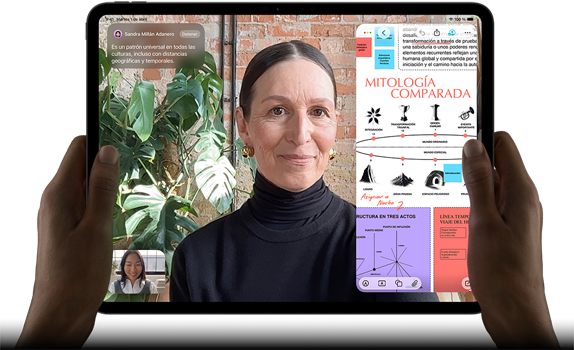 iPad Pro, parte delantera exterior, posición horizontal, color negro espacial, esquinas redondeadas, borde de la pantalla negro, dos manos sujetan los laterales izquierdo y derecho, la pantalla muestra una conversación de FaceTime con traducción en tiempo real y una app para aprender con una presentación sobre mitología comparada con texto, diagramas y notas escritas a mano
